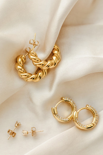 a flat lay of gold jewelry including croissant twisted hoops, chunky cz huggie earrings and 4mm cz stud earrings 