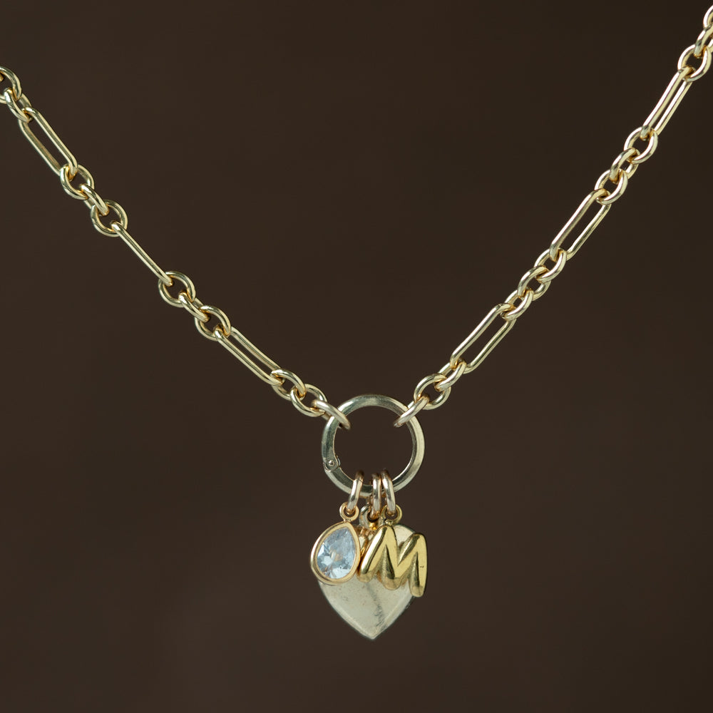 gold necklace with a carabiner in the center with gold charms dangling on a dark brown background 