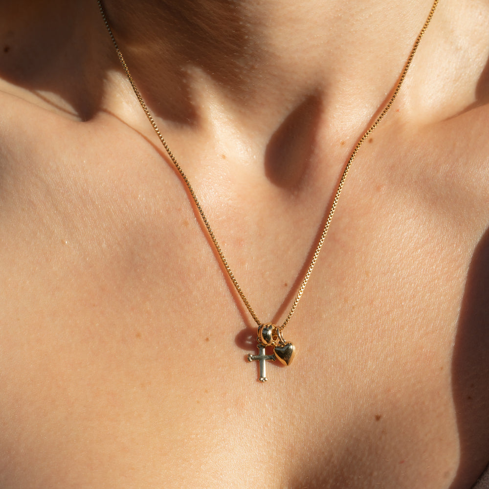 person wearing a gold box chain necklace with a heart charm, cross charm and gold bead spacer 