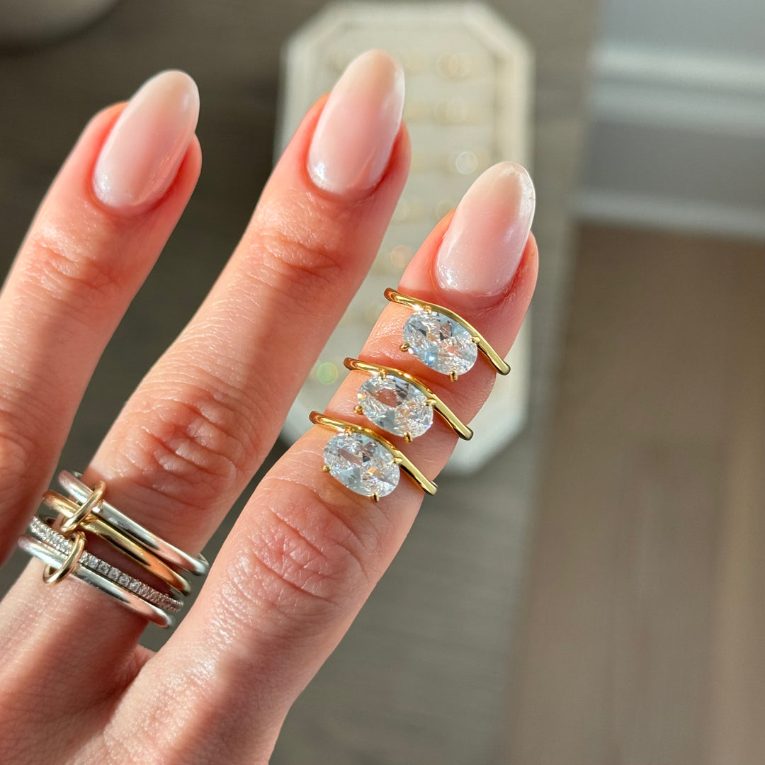 Hand wearing three gold rings with clear gemstones on a neutral background