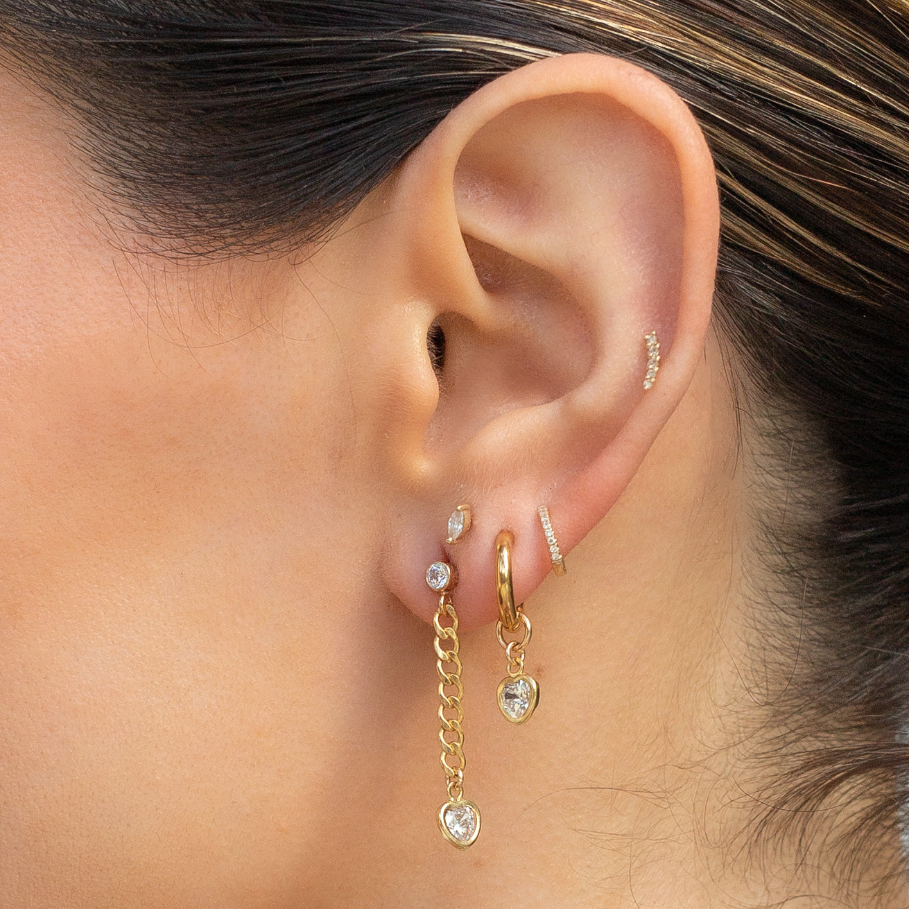 Ear with multiple earrings showing Round Cubic Zirconia Stud Earring with Hanging Curb Chain and CZ Heart Drop Charm Earring and Gold Hoop Huggies with CZ Heart drop charm